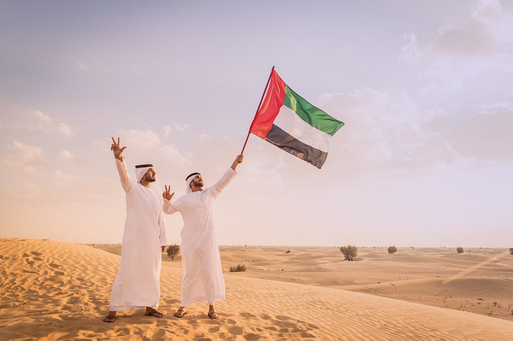 Image of 2 emarati men holding UAE flag and doing a victory sign using their hands in the desert.